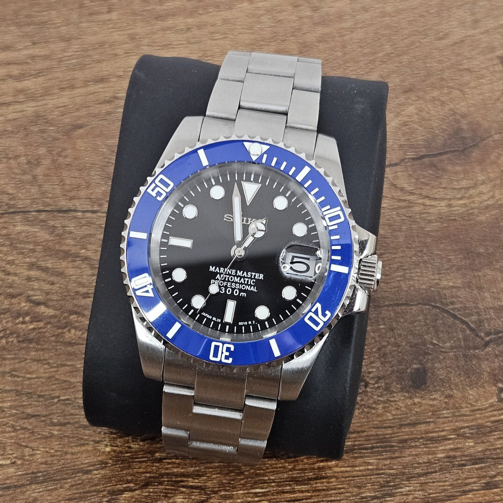 A steel watch with a blue ceramic bezel and a black sunburst dial, featuring silver hands and indexes, and a stainless steel bracelet.
