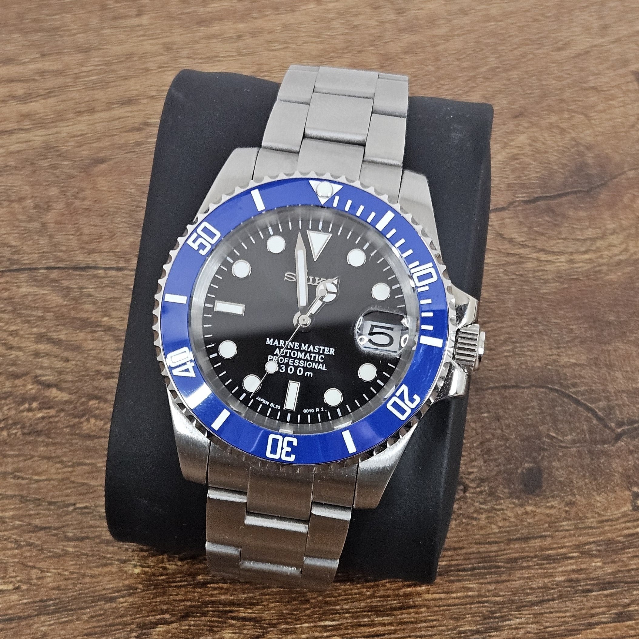 A steel watch with a blue ceramic bezel and a black sunburst dial, featuring silver hands and indexes, and a stainless steel bracelet.