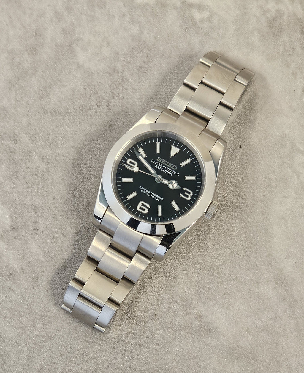 A Seiko Explorer Mod Watch with a black dial and a stainless steel bracelet, featuring a 36mm case with a black explorer style dial, on a textured grey background.