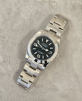 A Seiko Explorer Mod Watch with a black dial and a stainless steel bracelet, featuring a 36mm case with a black explorer style dial, on a textured grey background.