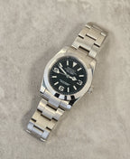 A Seiko Explorer Mod Watch with a black dial and a stainless steel bracelet, featuring a 36mm case with a black explorer style dial, on a textured grey background.