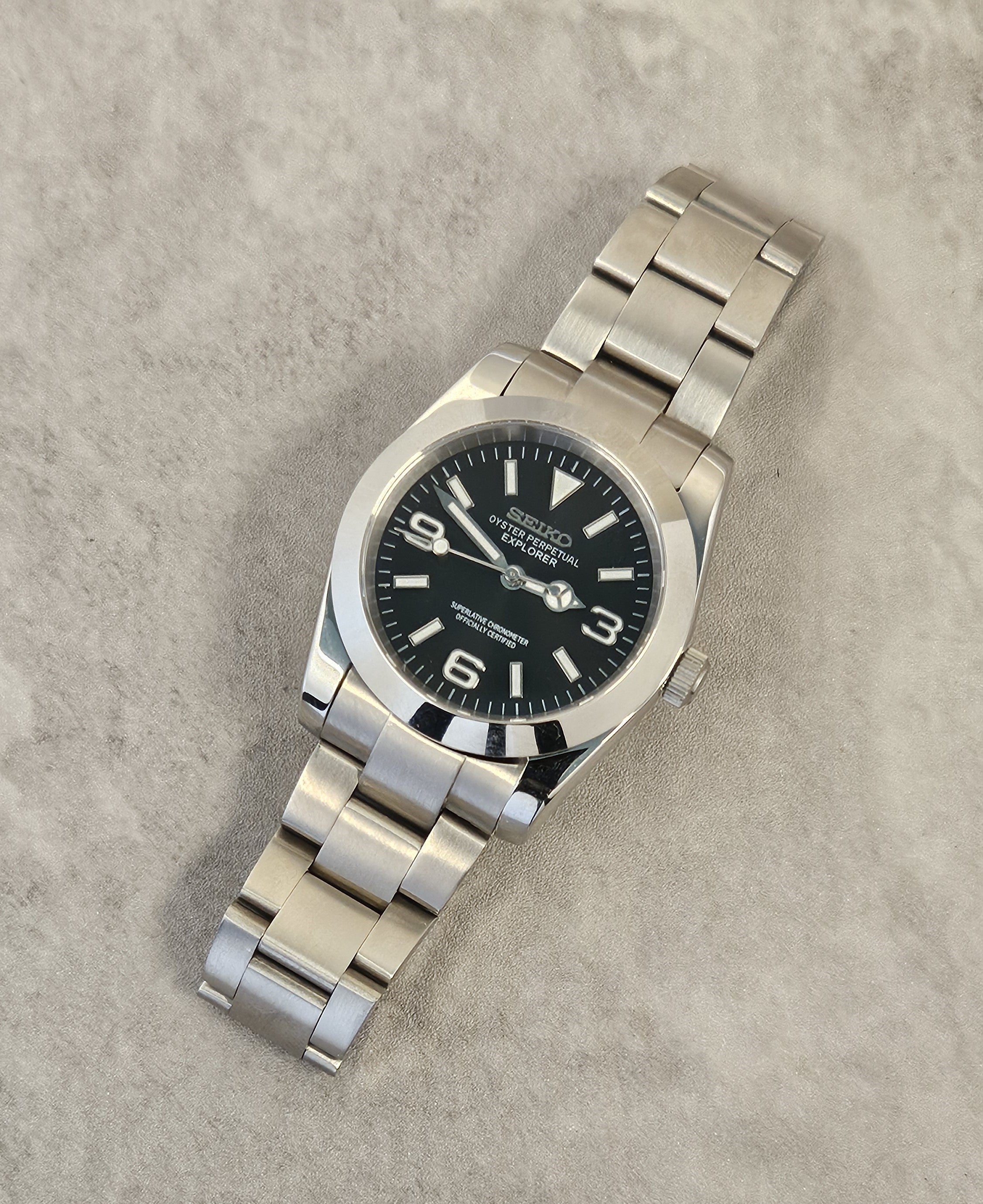 A Seiko Explorer Mod Watch with a black dial and a stainless steel bracelet, featuring a 36mm case with a black explorer style dial, on a textured grey background.