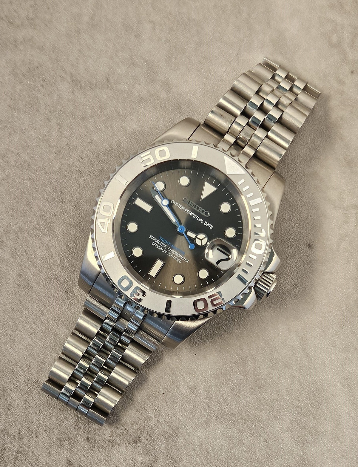 A luxurious silver-toned watch with a grey sunburst dial, luminous hands, a unidirectional rotating bezel, and a stainless steel bracelet on a textured background.