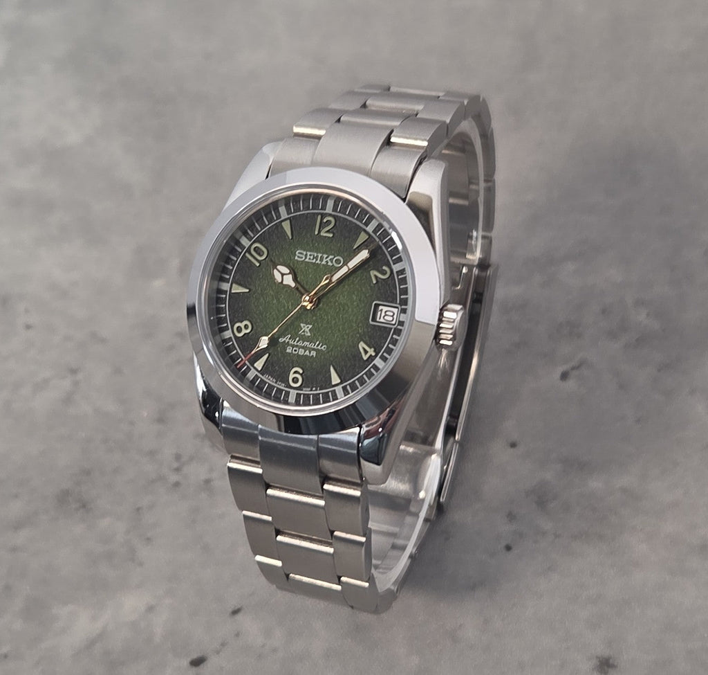 A Seiko stainless steel wristwatch with a green dial and silver link bracelet, photographed on a textured gray concrete surface.