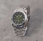 A Seiko stainless steel wristwatch with a green dial and silver link bracelet, photographed on a textured gray concrete surface.