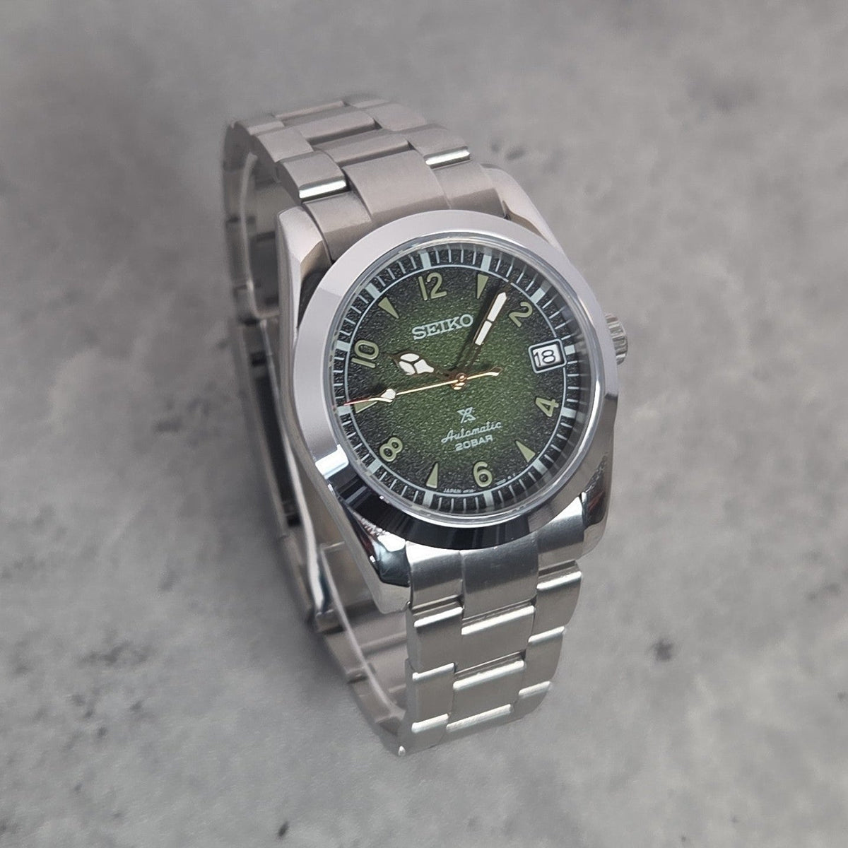 A Seiko stainless steel wristwatch with a green dial and silver link bracelet, photographed on a textured gray surface.