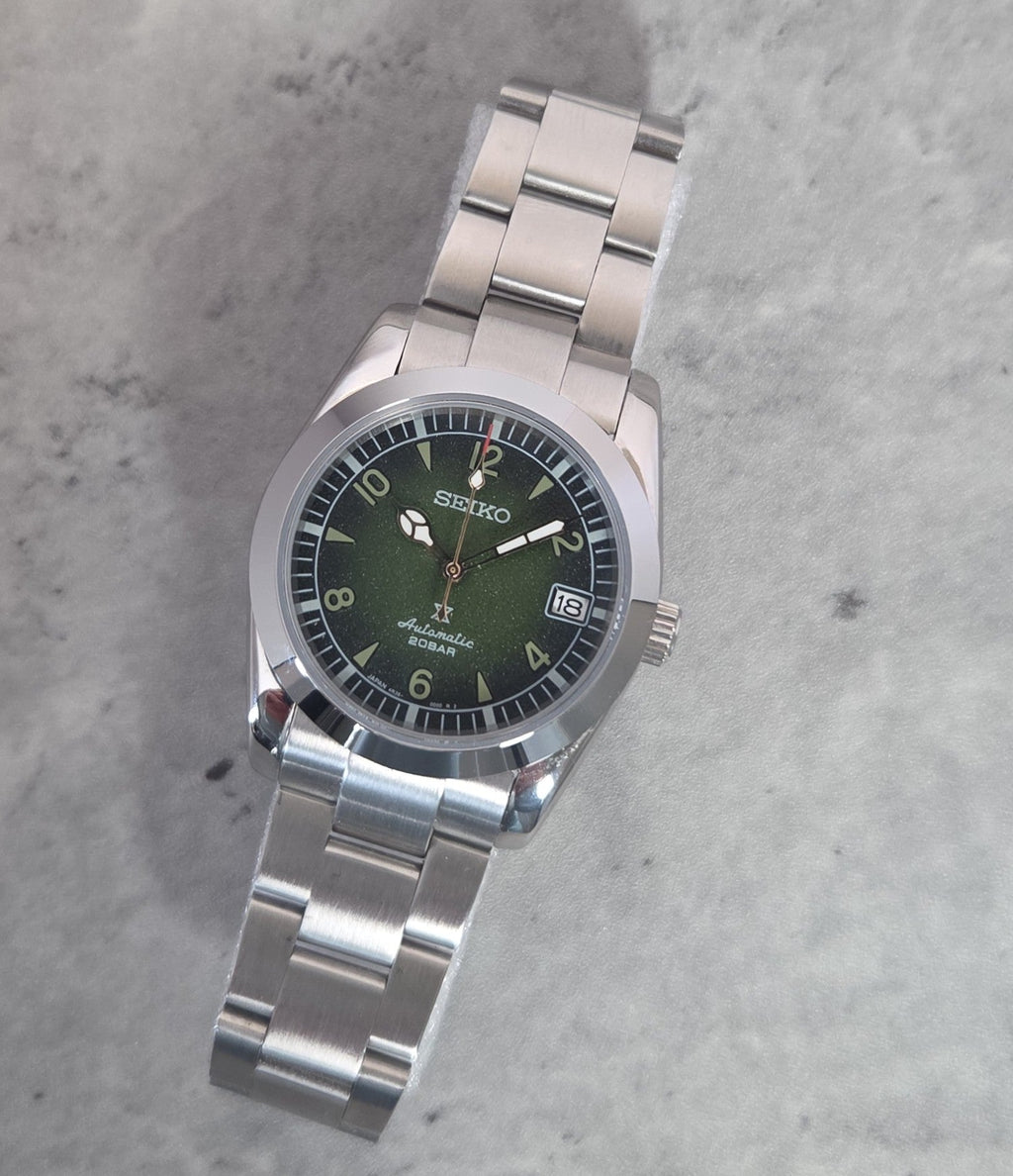 A Seiko Automatic Solar wristwatch with a green dial and a silver stainless steel link bracelet, photographed on a textured gray surface.