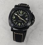 A Seiko GMT automatic watch with a black case, black dial, and black leather strap with light tan contrast stitching, photographed on a light gray marble surface.