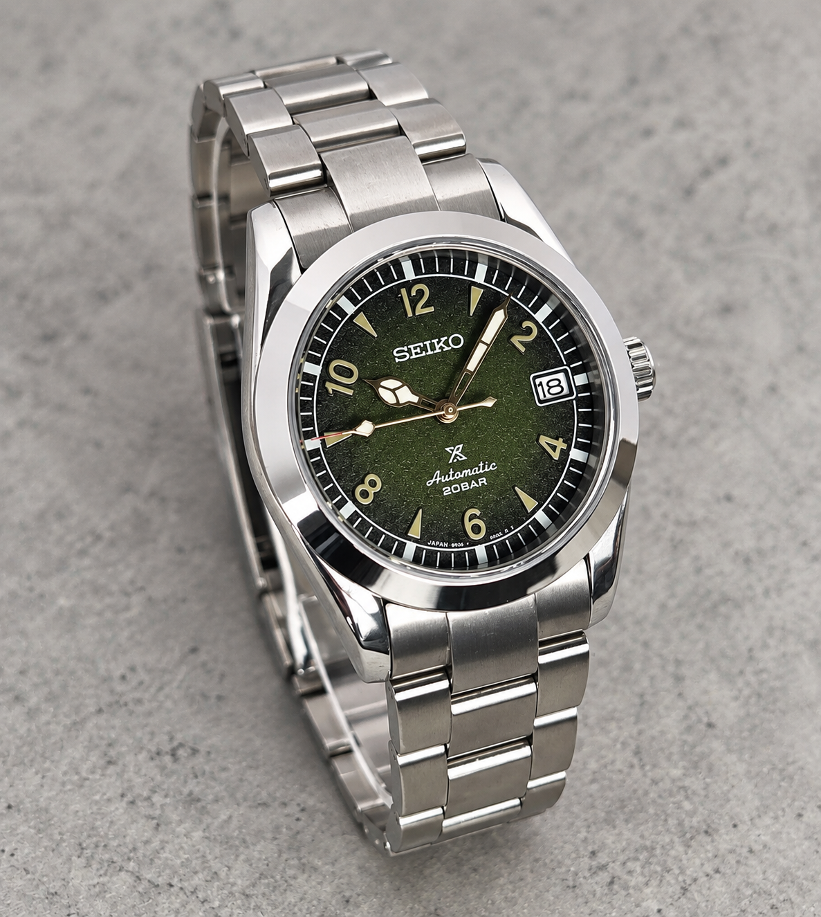 A Seiko Automatic 100m stainless steel watch with a dark olive green dial and matching green accents, displayed on a textured gray surface.