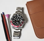 A GMT Master custom mod watch with a black dial, black and red bezel, and silver bracelet, displayed alongside a brown leather case, a cleaning brush, and a watch pen.