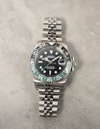 A GMT Master Spite Custom Mod Watch with a black dial, black and green bezel, and a stainless steel bracelet laid out next to a cup and pens.